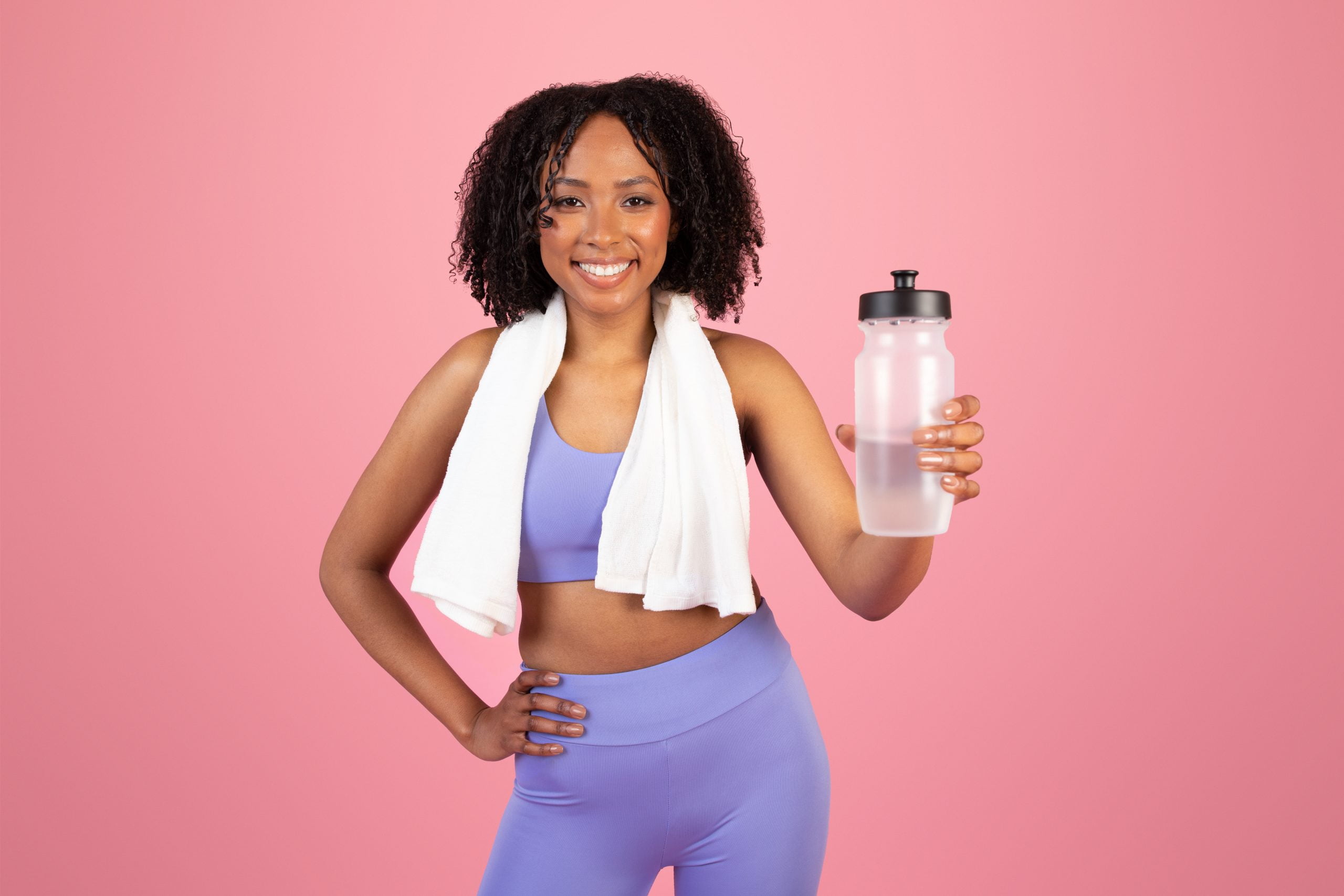 Positive young african american curly woman in sportswear with towel show bottle of water
