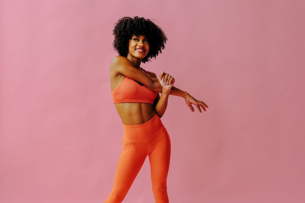 Smiling woman with curly hair in orange sportswear stretching her arm against a pink background - fit trainers, sustainable weight loss tips for busy women